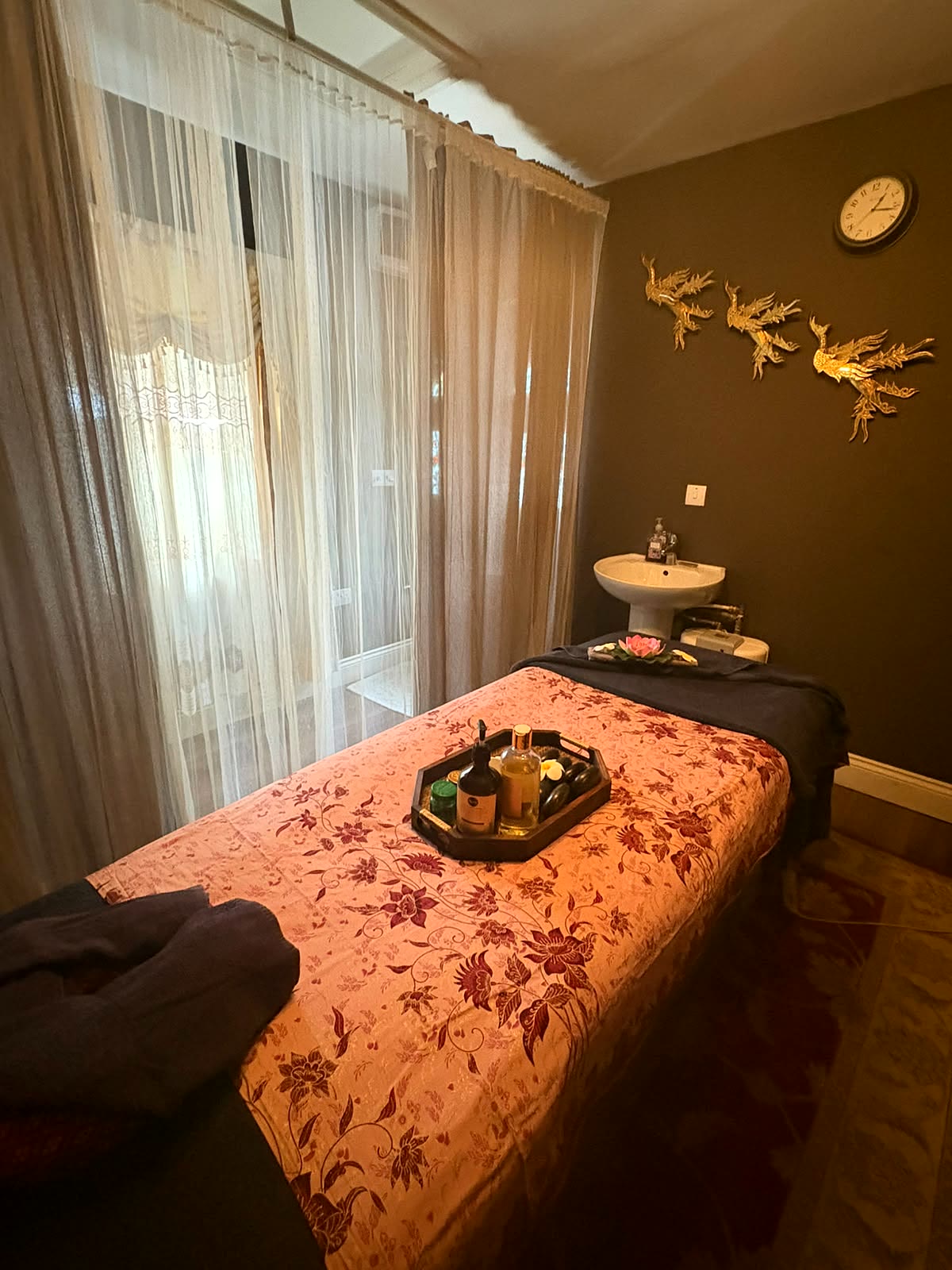 Sunlit sheer curtains beside the treatment bed, with the wash basin and gold phoenix wall art visible beyond