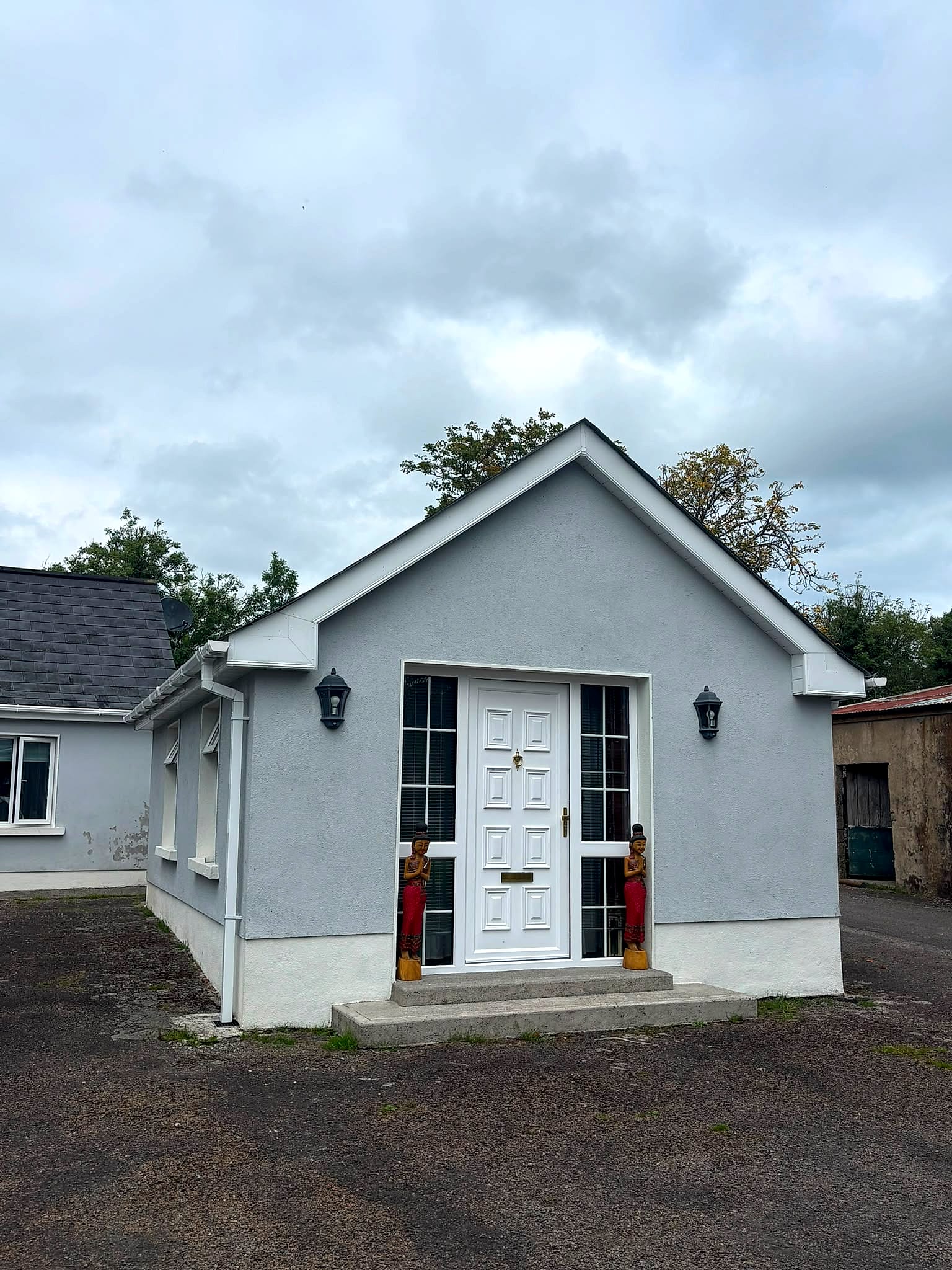 The grey-rendered studio building in Mohill, Co. Leitrim, with two carved wooden Thai greeting statues standing either side of the white front door
