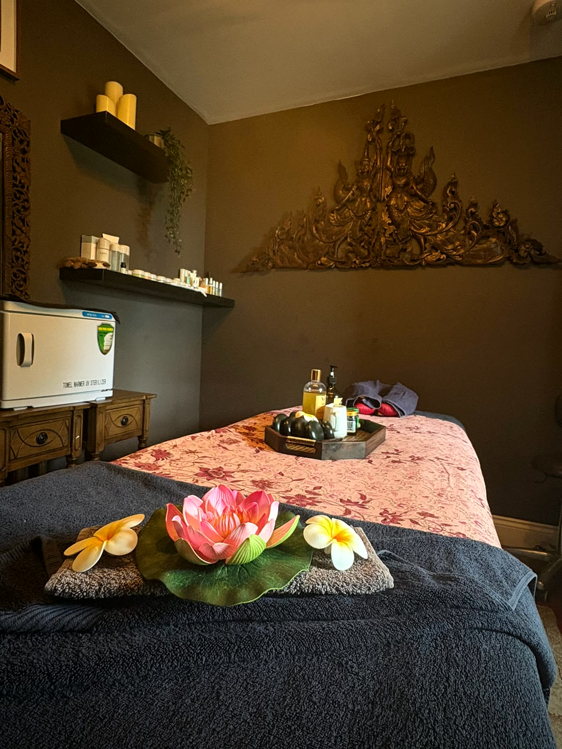 A wide view of the treatment room with a hand-carved gold Thai crown ornament above the bed, a shelf of oils and candles, a towel warmer, and a styled lotus flower arrangement in the foreground