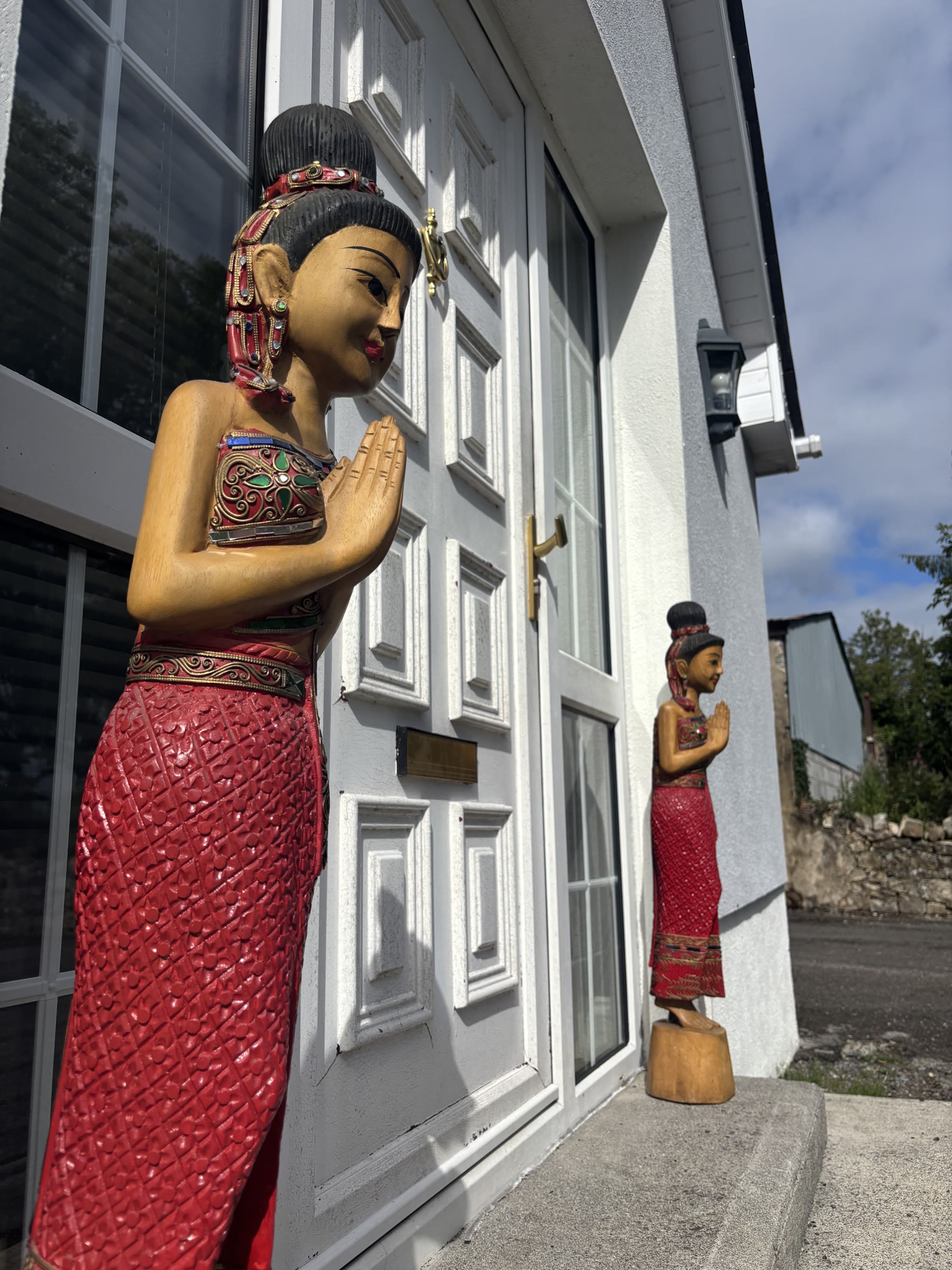 Two carved wooden Thai greeting statues, hands pressed together in a wai, standing either side of the front door